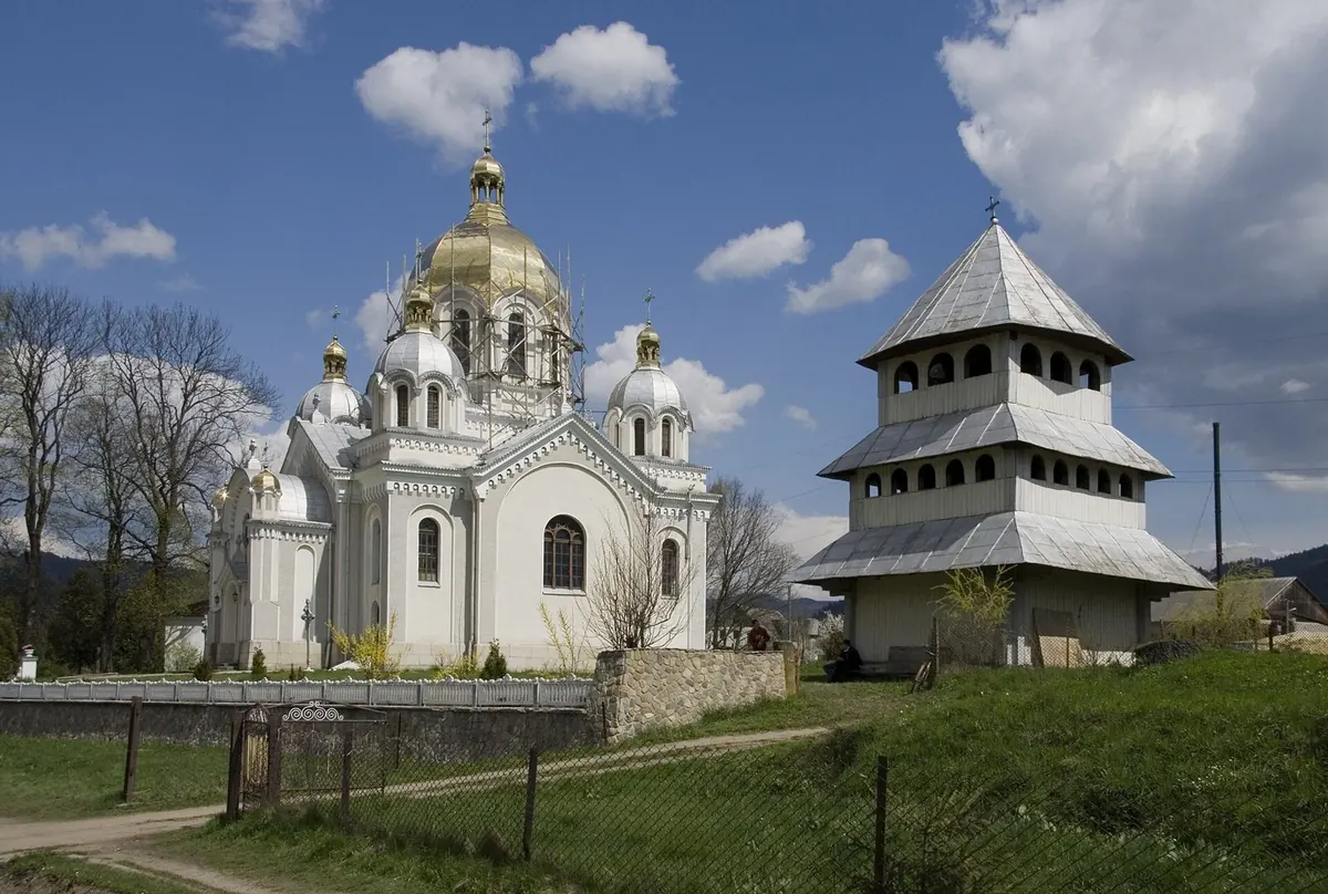 Біла мурована церква з позолоченими куполами і дерев'яна дзвіниця під синім небом