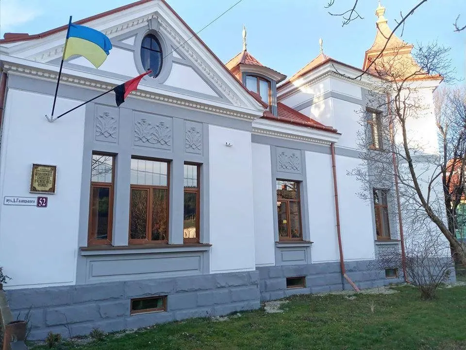 Білий будинок музею з різьбленим фронтоном і великими вікнами, на фасаді прапори