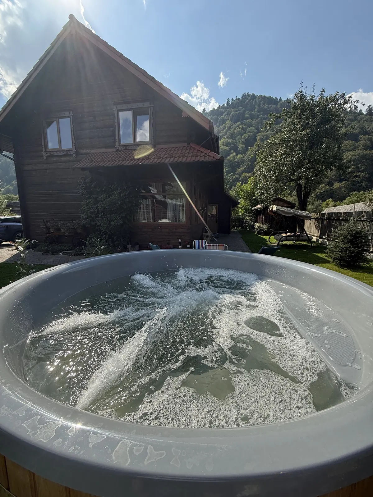 Чан з водою, що вирує, перед дерев'яною двоповерховою хатою з червоним дахом, позаду зелена гора і сонце