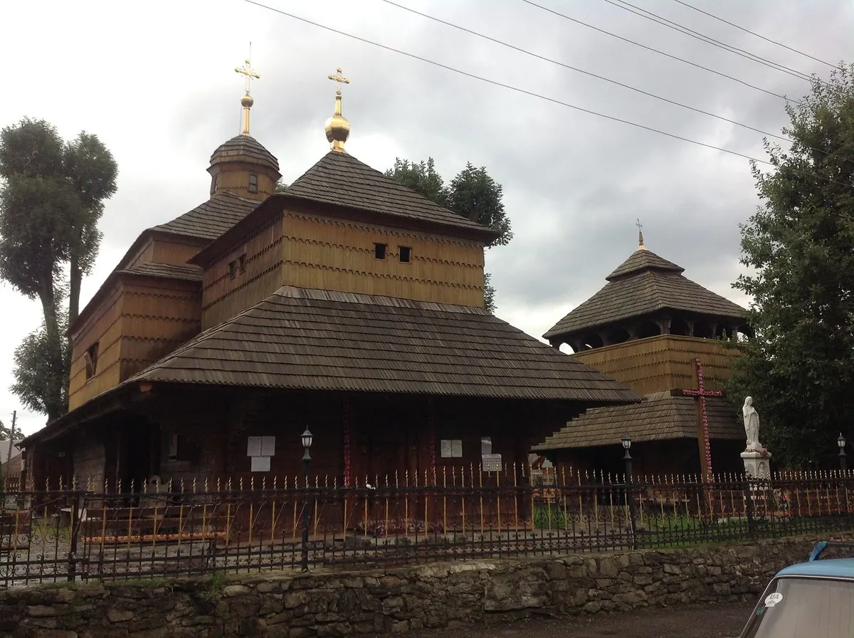 Дерев'яна бойківська церква з ґонтовими дахами й двома маківками, поруч дерев'яна дзвіниця за кованою огорожею
