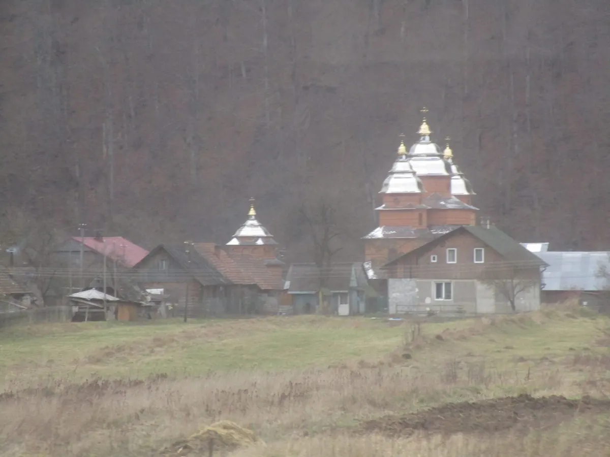 Дерев'яна церква з позолоченими верхами серед хат біля підніжжя лісистої гори, зимовий день