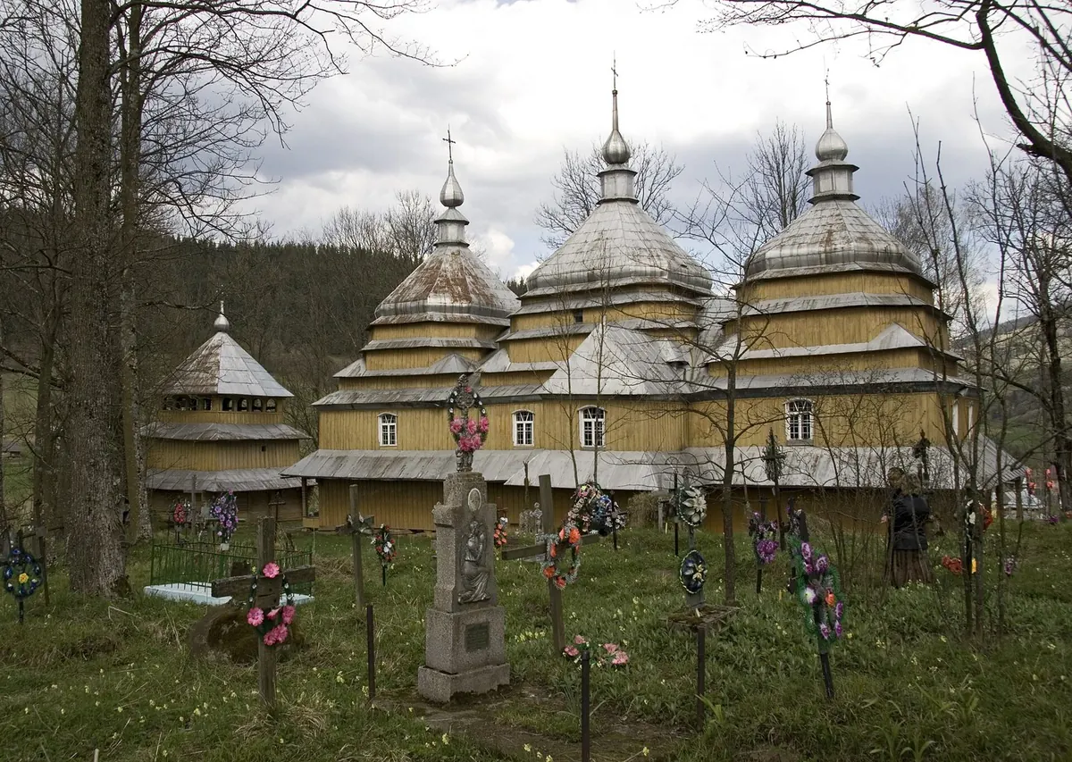 Дерев'яна церква з трьома верхами й окрема дзвіниця на пагорбі, перед ними цвинтар з хрестами