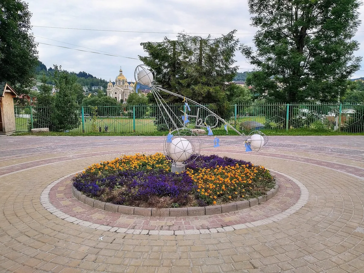 Металевий арт-об'єкт із кулями на клумбі з фіалками посеред бруківки, вдалині церква з позолоченими куполами