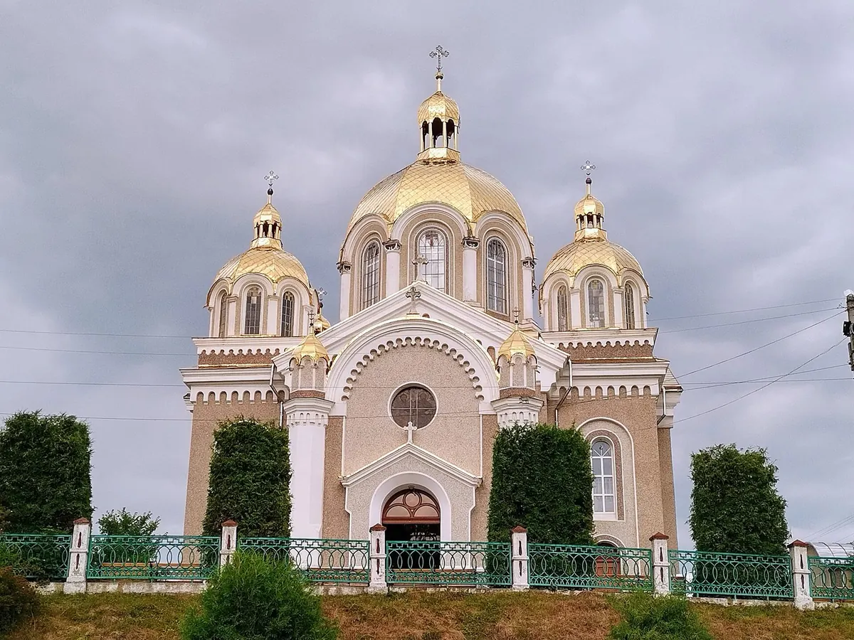 Мурована церква Петра і Павла з позолоченими куполами за зеленою огорожею, хмарне небо