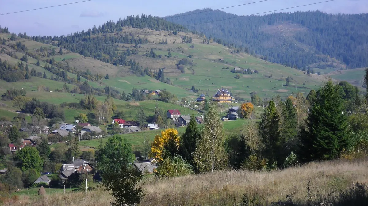 Осінні пагорби над селом, між хатами дерев'яна будівля, позаду лісистий хребет