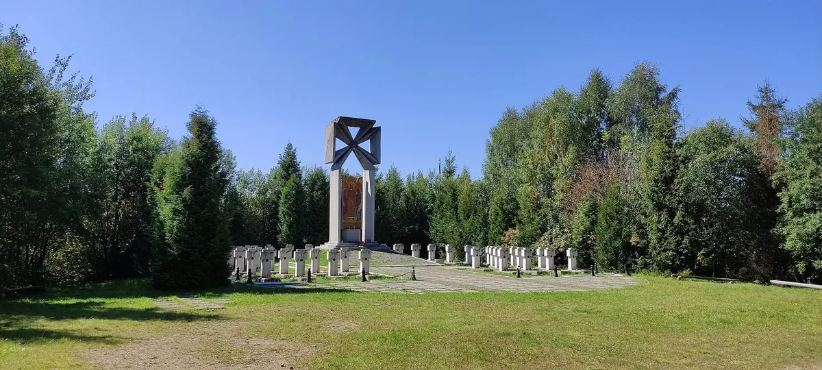 Сонячна галявина серед ялин і беріз: два ряди білих хрестів обабіч високого пам'ятника з хрестом угорі та іконою