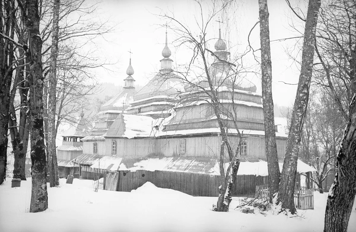 Старе чорно-біле фото: дерев'яна церква з трьома верхами взимку серед високих дерев, усе в снігу