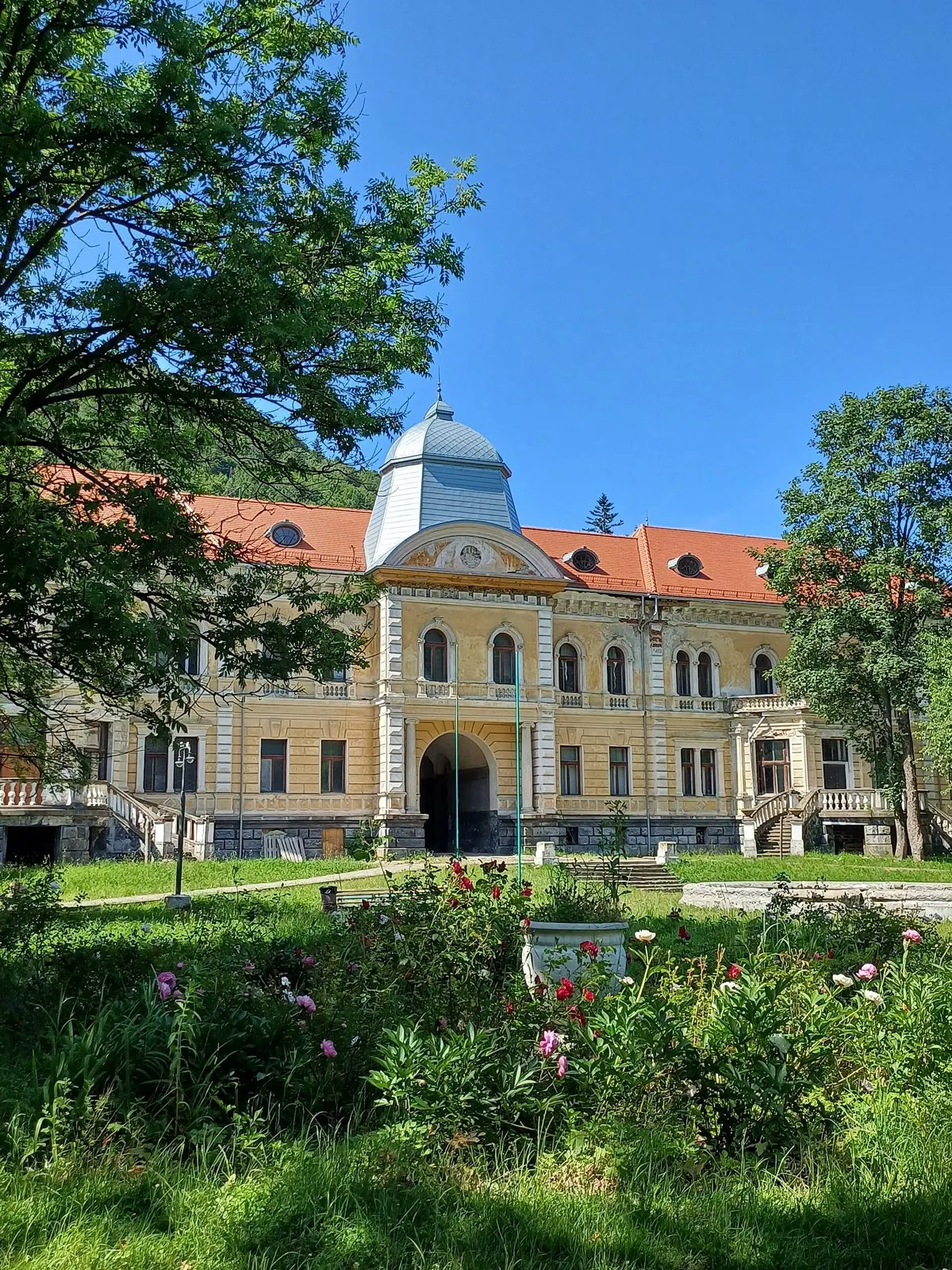 Світлий двоповерховий палац з червоним дахом і баштою, перед ним квітник і дерева старого парку